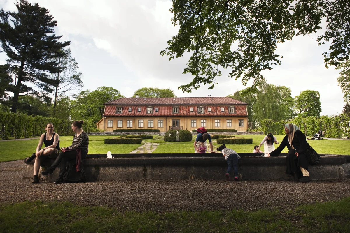 People sitting by an fountain in front of a garden and an old building called Tøyen Manor